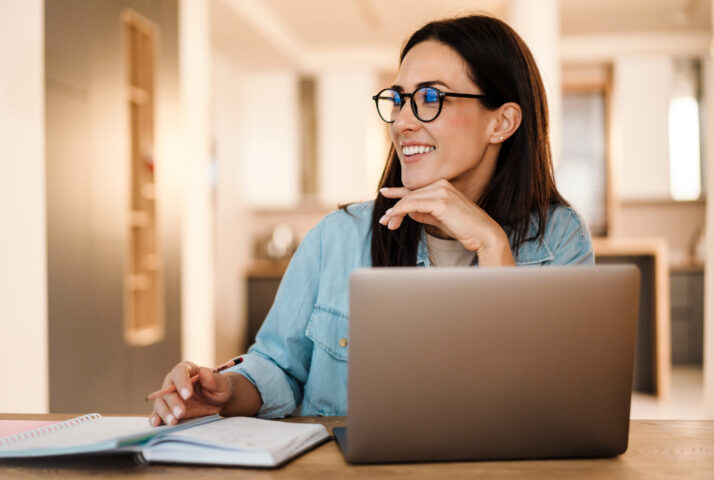 Happy charming woman writing down notes while working with laptop at home